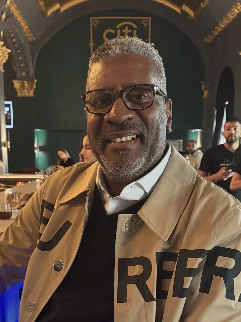 Smiling man in glasses and a tan jacket at an evening event in an ornate hall